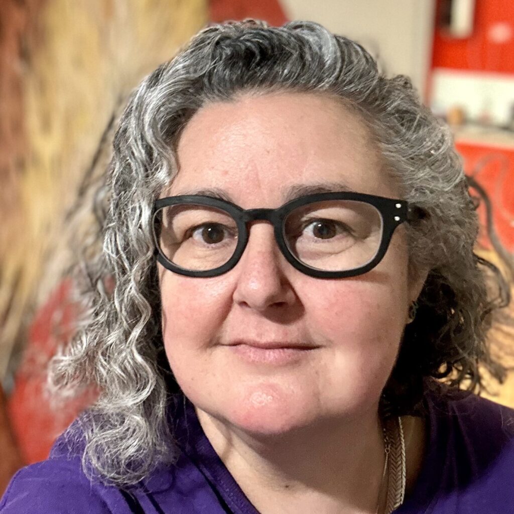 Woman with curly salt and pepper curly hair and black rimmed glasses in a purple shirt