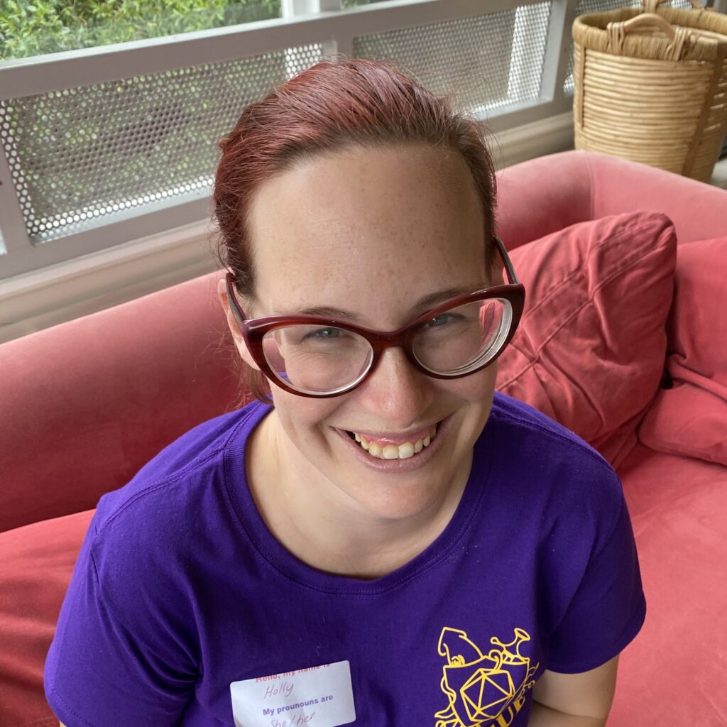 Smiling red haired woman on a red couch, with a purple t-shirt and red glasses