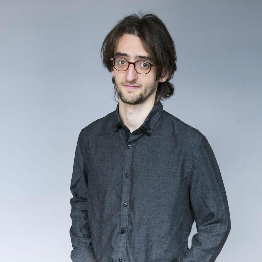 skinny scruffy white guy in a dark shirt with glasses, a quizzical expression and patchy facial hair