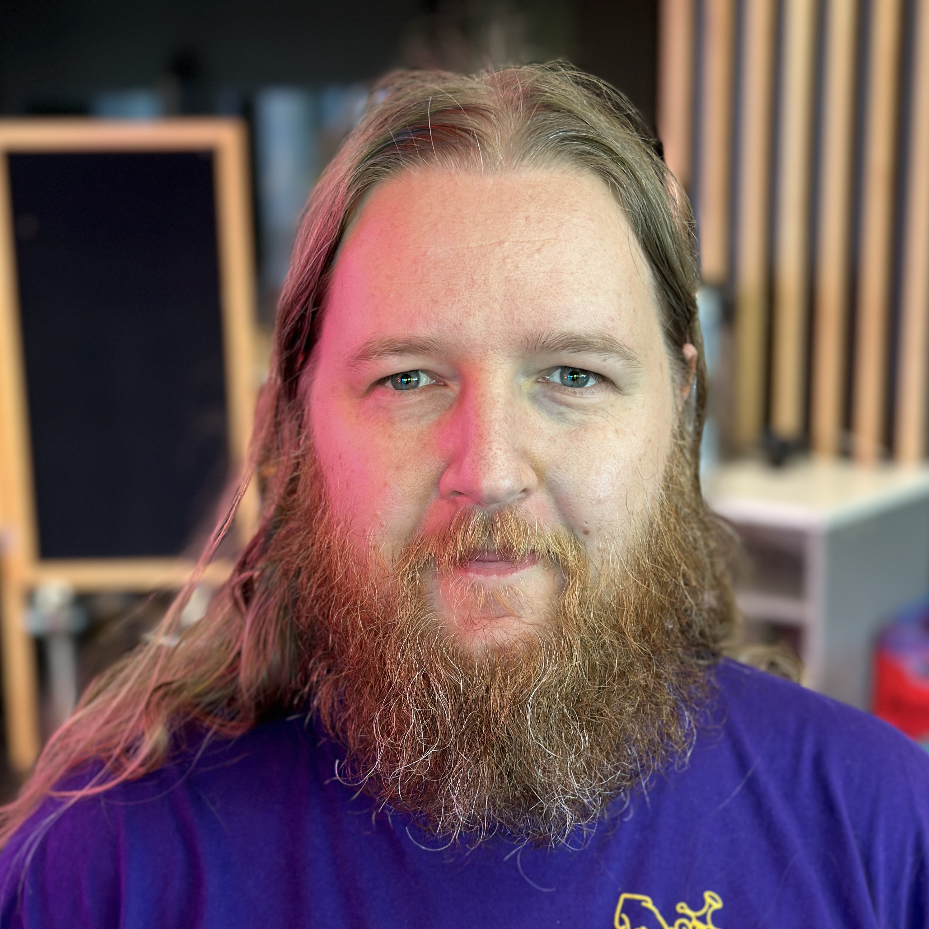 a person with long red hair, a bushy red beard, a purple t-shirt and an enigmatic smile.