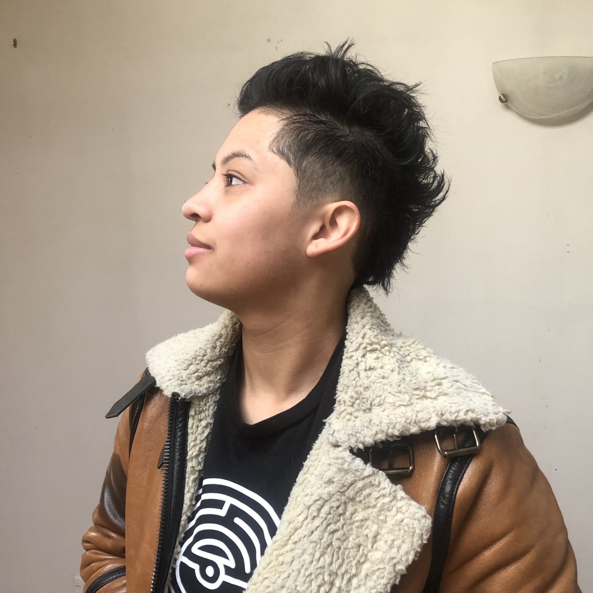 young person with short dark spiky hair in profile, wearing a shearling leather jacket and black t-shirt against a neutral wall